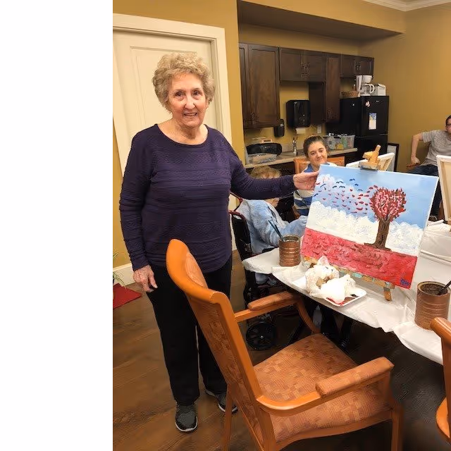 An elderly woman standing and smiling while holding a painting of a tree with a heart-shaped canopy and birds flying in a blue sky with white and red layers below. Behind her, two other women are seated at a table in a room with wooden cabinets and a refrigerator, engaged in an art activity.