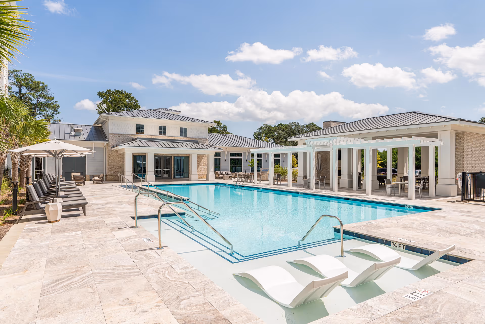 Outdoor swimming pool area at Overture West Ashley with lounge chairs, umbrellas, and a pergola-covered seating area. The pool has built-in lounge chairs in the shallow end and is surrounded by a tiled deck. The building in the background has a modern design with large windows and a metal roof under a partly cloudy blue sky.