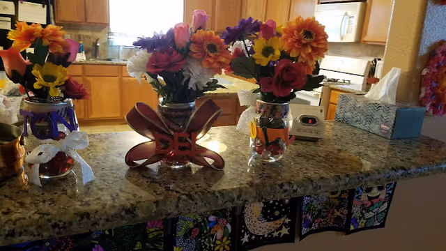 A kitchen countertop with three glass vases filled with colorful artificial flowers. Each vase is decorated with ribbons and letters. Behind the counter, there is a kitchen area with wooden cabinets, a microwave, a stove, and a tissue box on the counter. The front of the counter is decorated with colorful patterned paper.