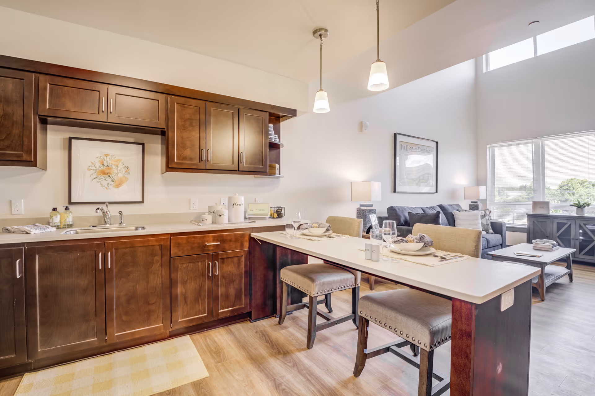 A bright and modern open-concept living space featuring a kitchen area with dark wood cabinets, a sink, and countertop. Adjacent to the kitchen is a dining area with a white table set for two with plates, glasses, and napkins. Beyond the dining area is a living room with a gray sofa, coffee table, sideboard, and large windows letting in natural light.