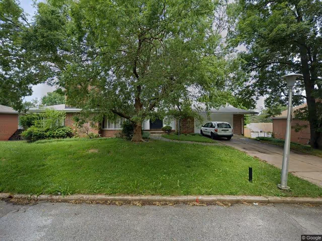 A single-story brick building partially obscured by a large leafy tree in the front yard. There is a driveway with a white car parked on it, a grassy lawn, and a streetlamp on the right side near the sidewalk. The scene appears to be a residential or facility front exterior view with surrounding greenery.
