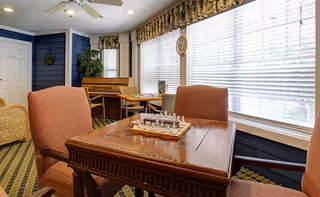 Sunlit common room with a wooden table and upholstered chairs by large windows and a chess set on the table.