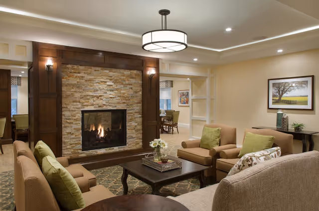 Cozy living room with a stone fireplace surrounded by dark wood paneling, beige walls, and recessed lighting. The room features comfortable beige and brown upholstered chairs and sofas with green and patterned cushions, a dark wooden coffee table with a flower arrangement, and a framed landscape painting on the wall. In the background, a dining area with a wooden table and chairs is visible.