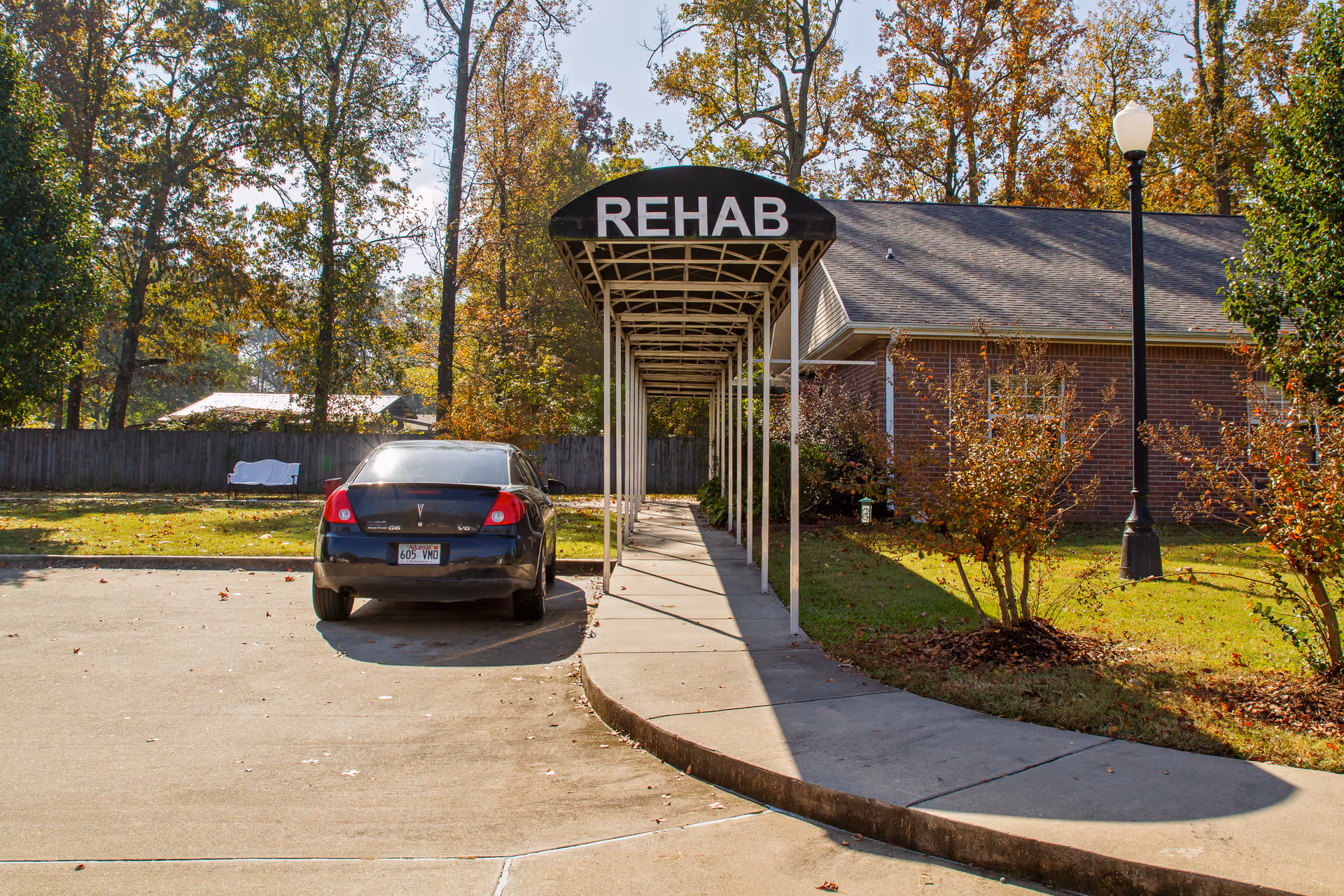 Outdoor view of a covered walkway labeled 'REHAB' leading to a brick building, with a black car parked nearby and trees with autumn foliage in the background.
