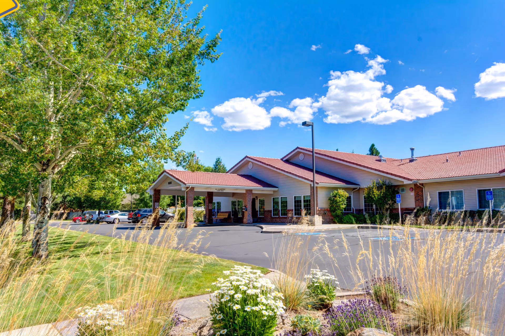 Exterior view of Aspen Ridge Memory Care facility on a sunny day with a clear blue sky and scattered clouds. The building has a red-tiled roof and light-colored walls with brick accents. There is a covered entrance with columns, a parking lot with several cars, and landscaped greenery including trees, tall grasses, and flowering plants in the foreground.