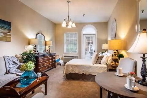 A cozy senior living bedroom featuring a bed with white bedding, a wooden dresser with a mirror, a small round table with two cups and a lamp, a comfortable armchair, and a sofa with a blanket and cushions. The room is warmly lit with lamps and a ceiling light, and has a window and a glass door leading outside.