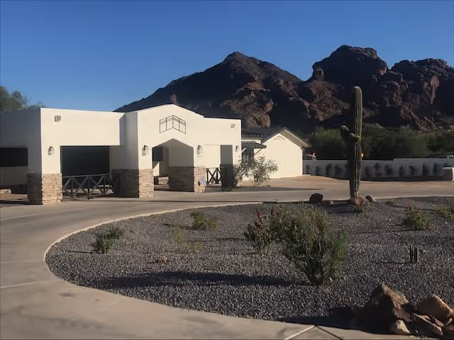 Front entrance of a low white assisted living building with a circular driveway, desert landscaping, and rocky hills in the background.
