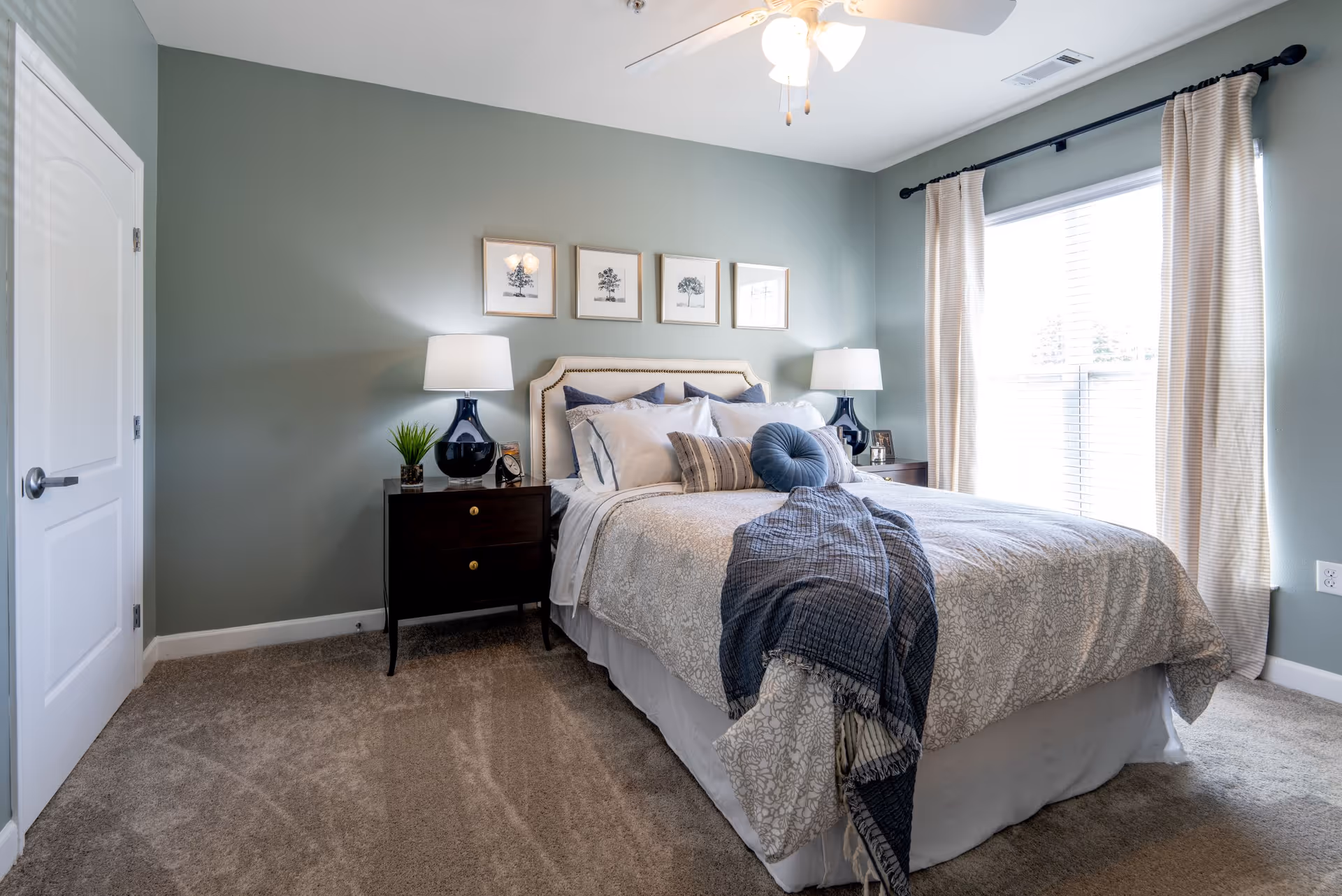 A neatly made bed with multiple pillows and a throw blanket in a bedroom with light green walls. There are two dark wooden nightstands on either side of the bed, each with a lamp and decorative items. Above the bed are four framed botanical prints. A large window with beige curtains allows natural light into the room, and a ceiling fan with lights is visible above.