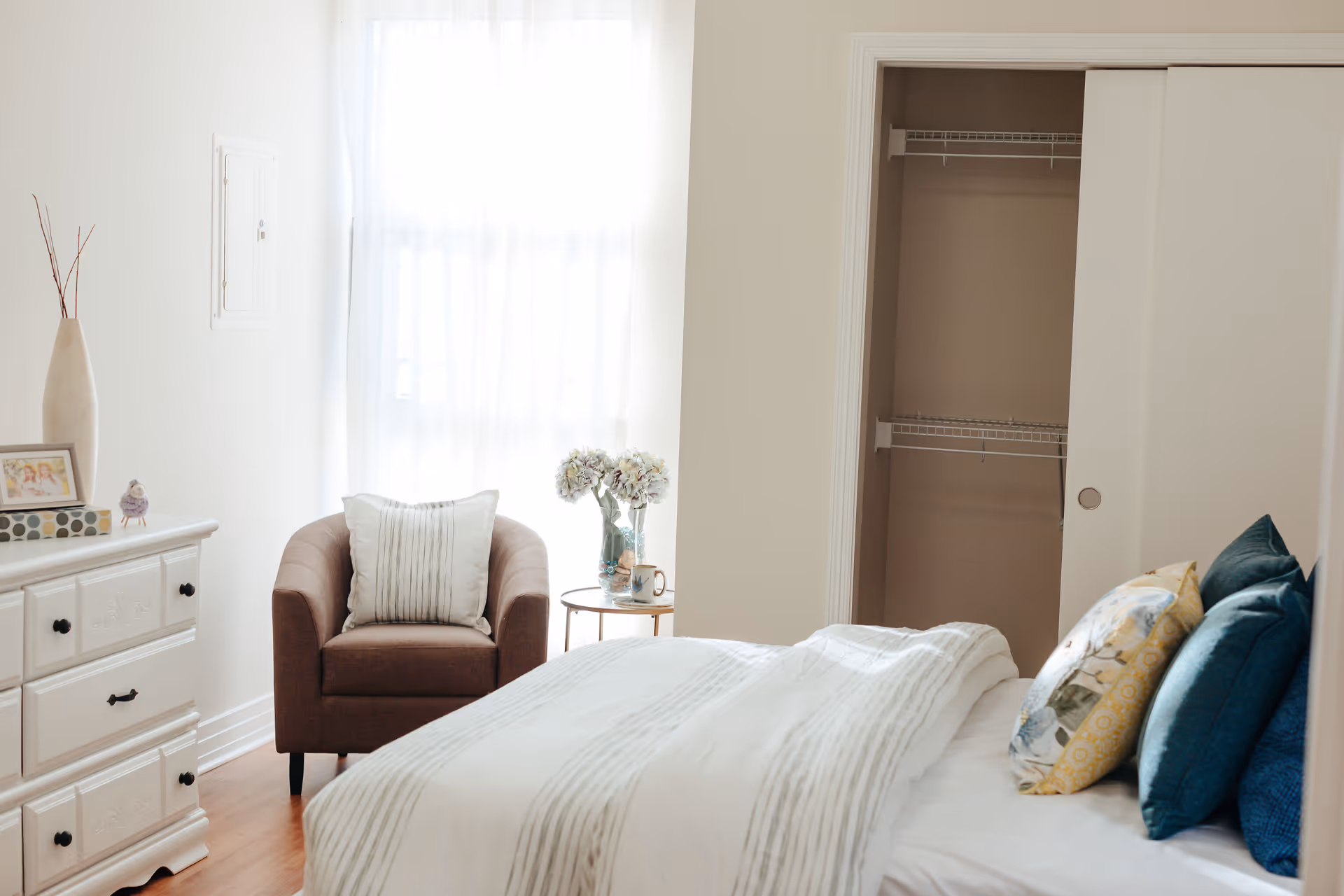 A bright bedroom with a neatly made bed featuring multiple pillows, a brown armchair with a white cushion, a small round side table with a vase of flowers and a mug, a white dresser with decorative items, and an open closet with wire shelving.