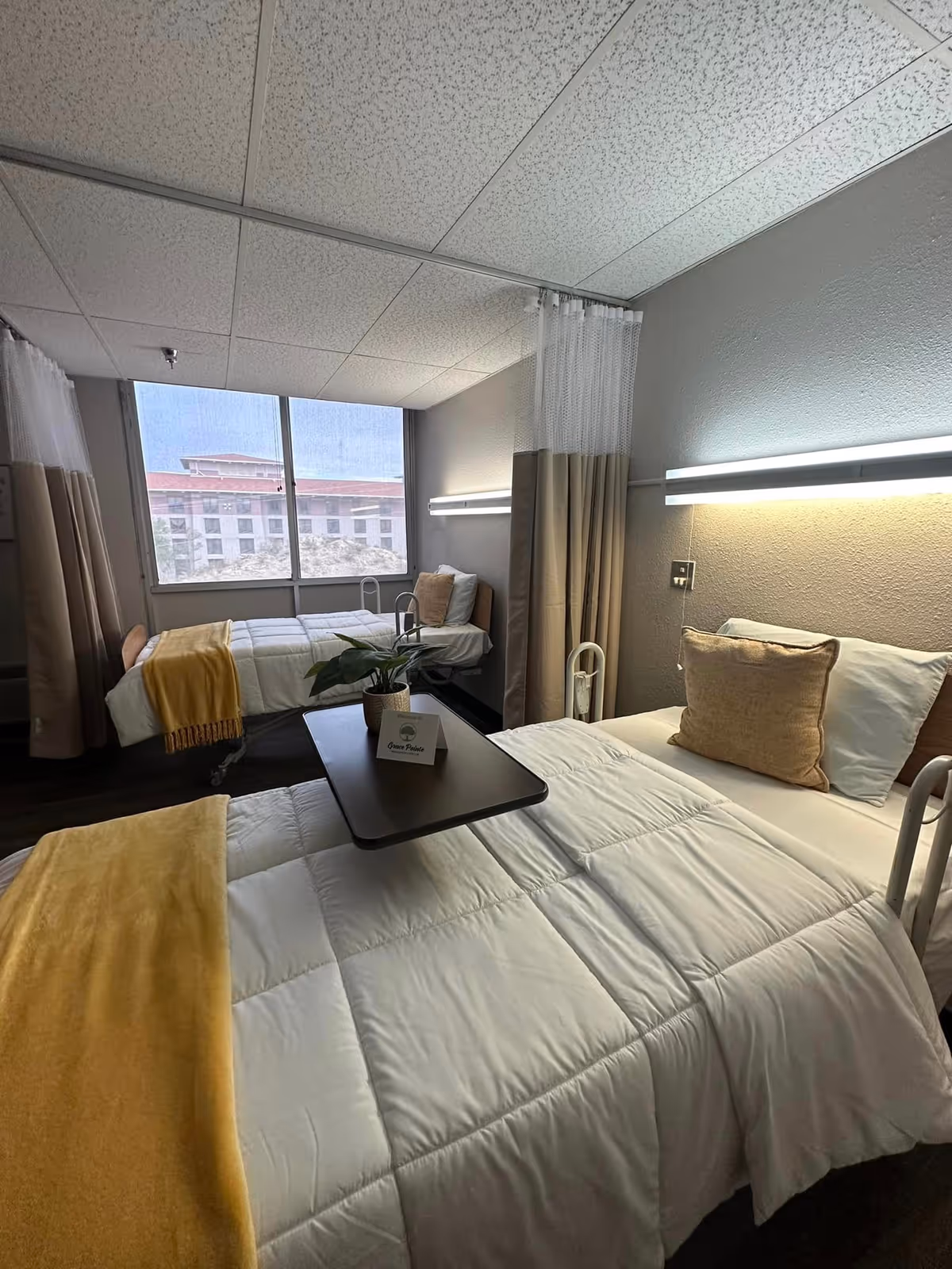 Two hospital-style beds in a shared senior living room separated by a privacy curtain, with white bedding, yellow throws, an overbed table with a plant, and a large window.