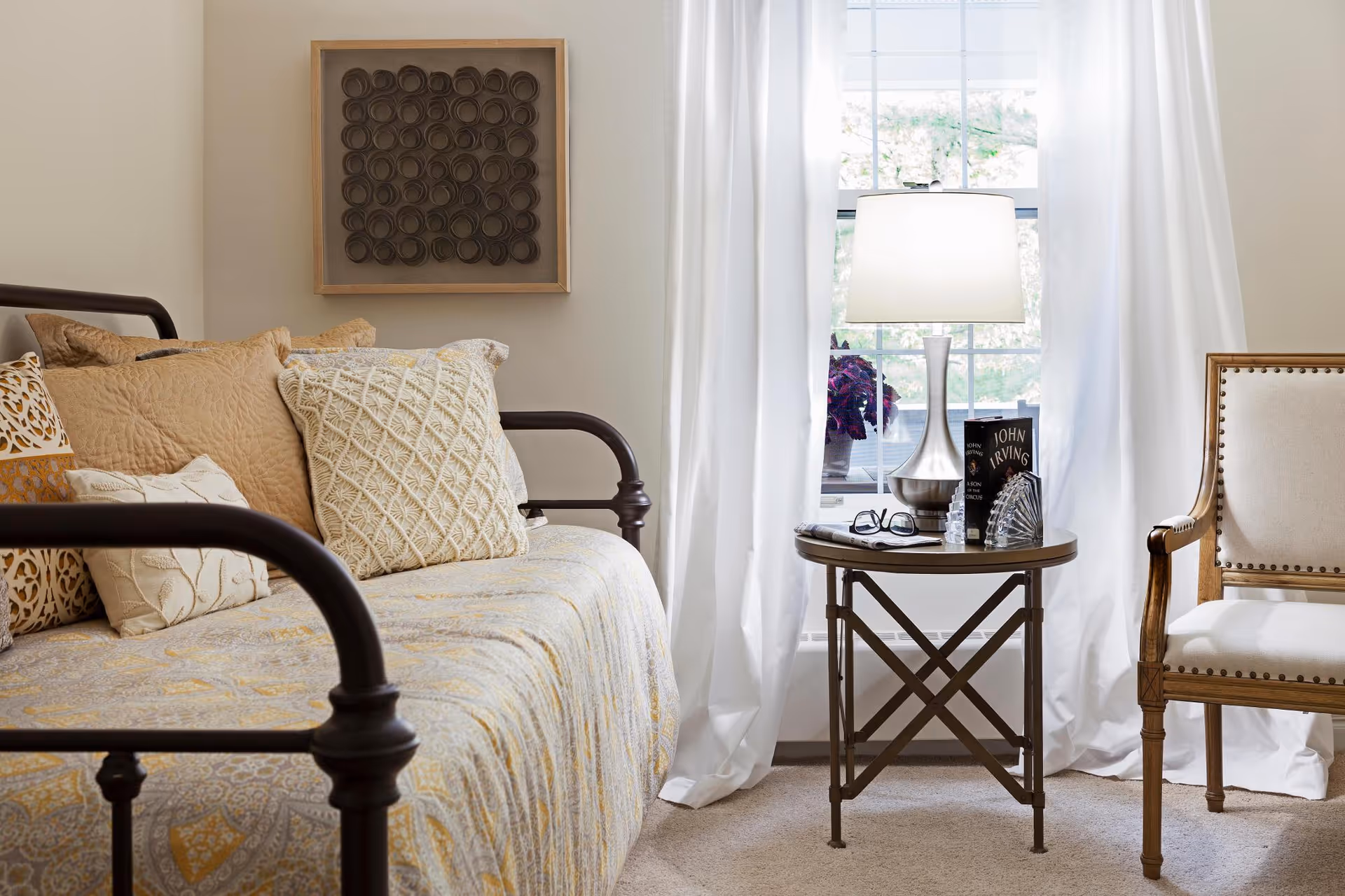 A cozy bedroom corner with a metal-framed daybed covered in patterned pillows, a round side table holding a lamp and books, and a window with white curtains.