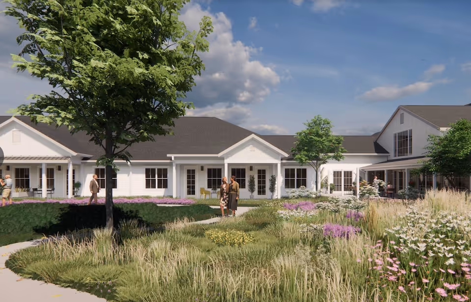 Exterior view of Cedarhurst Senior Living of Topeka showing a white building with multiple windows and a dark roof, surrounded by landscaped gardens with green grass, flowering plants, and trees under a partly cloudy sky. Several people are walking and standing on the pathways near the building.