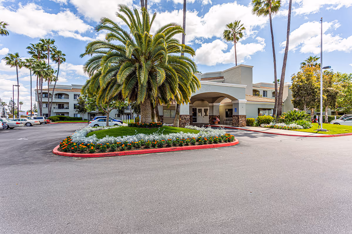 Front entrance of a senior living facility with a circular driveway, palm trees and landscaped flowerbeds.