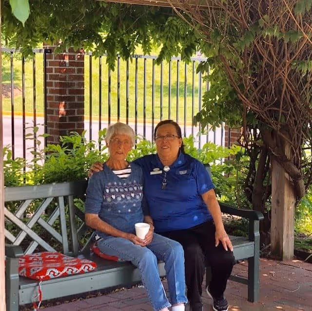 Two women sitting on a green wooden bench under a leafy pergola. One woman is elderly, wearing a blue sweater and jeans, holding a white cup. The other woman, wearing a blue polo shirt and black pants, has her arm around the elderly woman. There is a red and white patterned blanket on the bench beside them, and a black metal fence and greenery in the background.