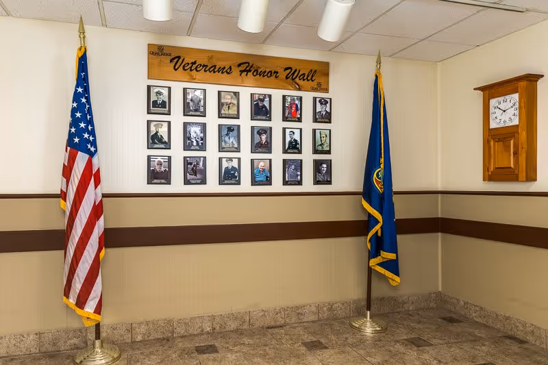 Interior corner of a room with a Veterans Honor Wall displaying framed photos of veterans. Two flags, the American flag and another flag, stand on either side of the wall. A wooden clock is mounted on the adjacent wall.