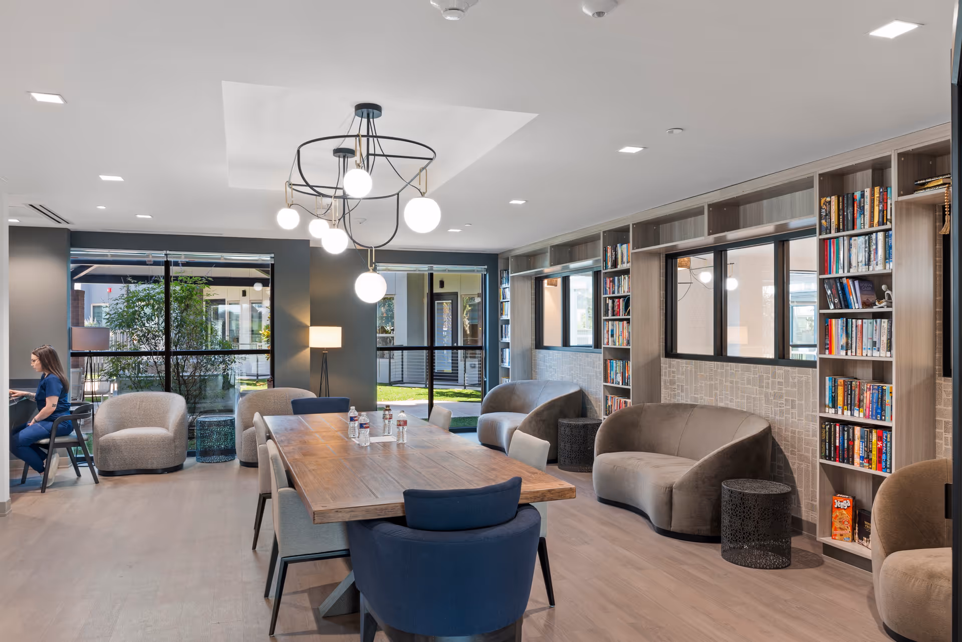 A modern common area with a large wooden table surrounded by chairs, several gray armchairs, built-in bookshelves filled with books, large windows letting in natural light, and a contemporary chandelier hanging from the ceiling. A person is seated at a desk in the corner near the windows.