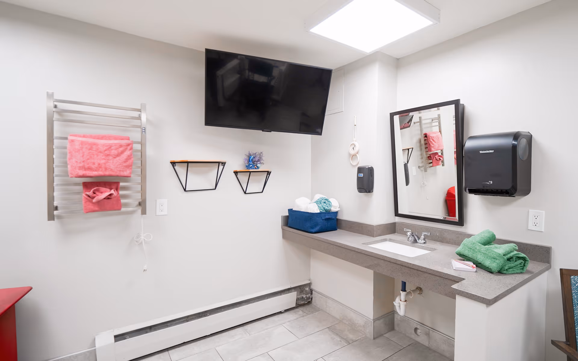 A clean bathroom with a wall-mounted flat screen TV, a towel rack holding pink towels, two small wall shelves, a countertop with a sink, a mirror, a black paper towel dispenser, and green towels on the counter. The floor is tiled and the walls are painted white.