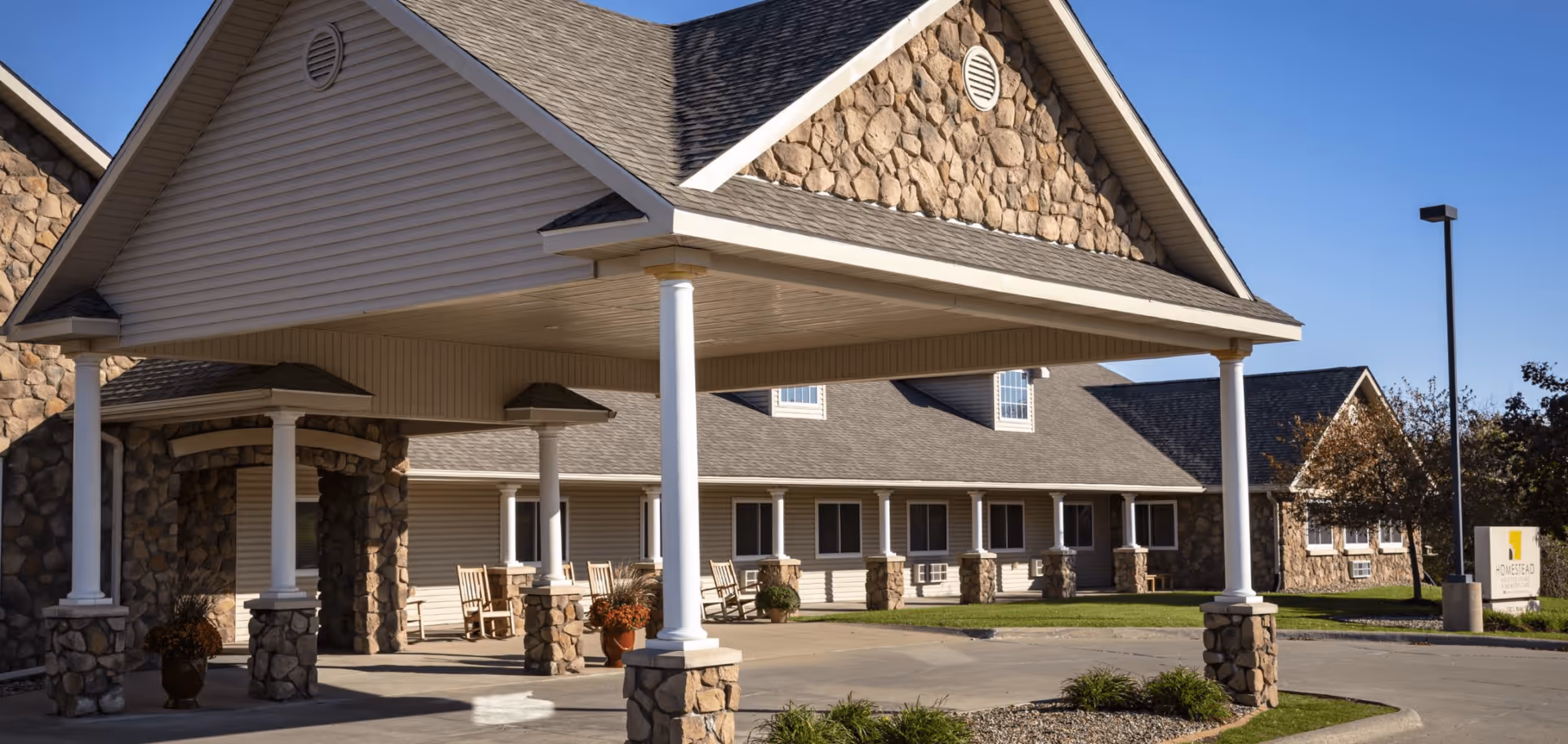 Exterior view of Homestead Assisted Living & Memory Care of Oskaloosa showing a covered entrance with stone pillars and rocking chairs on the porch under a clear blue sky.