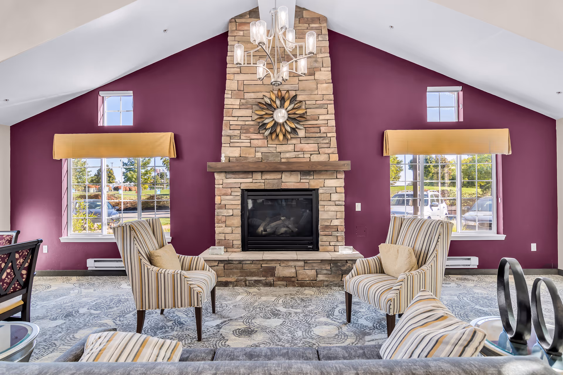 Sunlit common living room with a central stone fireplace flanked by windows and striped armchairs.