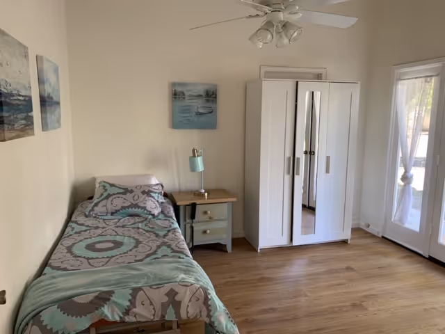 A simple bedroom with a single bed covered in a patterned bedspread in shades of teal, gray, and white. Next to the bed is a small nightstand with a teal lamp. On the wall above the bed are two framed pictures. Across from the bed is a white wardrobe with mirrored doors. To the right, there is a glass door with sheer curtains allowing natural light to enter the room. The floor is wooden, and the walls are painted light beige.