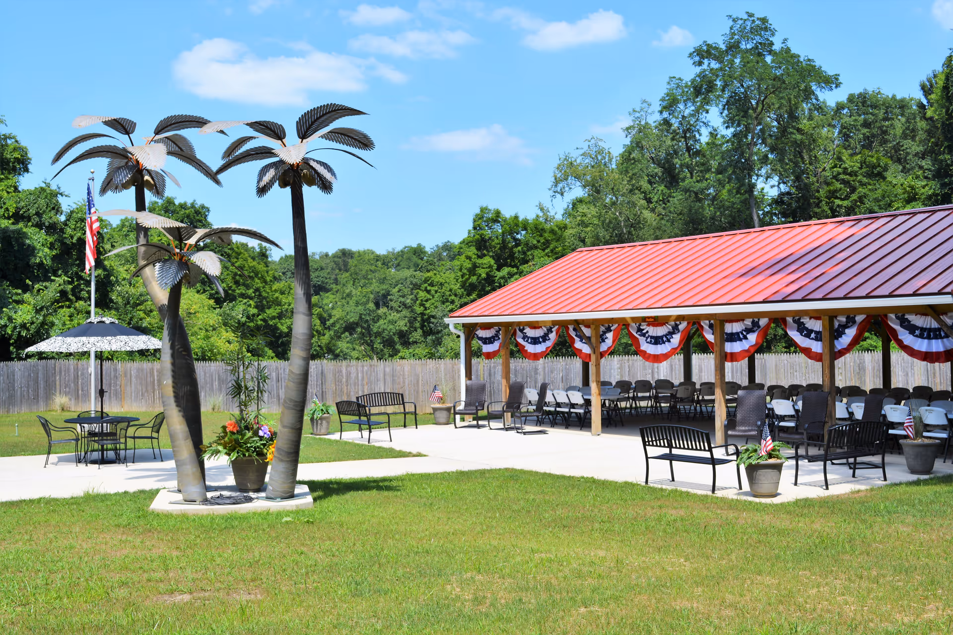 Outdoor area with a covered pavilion decorated with red, white, and blue bunting. Several chairs and benches are arranged under the pavilion and on the concrete patio. There are three metal palm tree sculptures and a table with an umbrella nearby. The area is surrounded by a wooden fence and trees in the background under a clear blue sky.