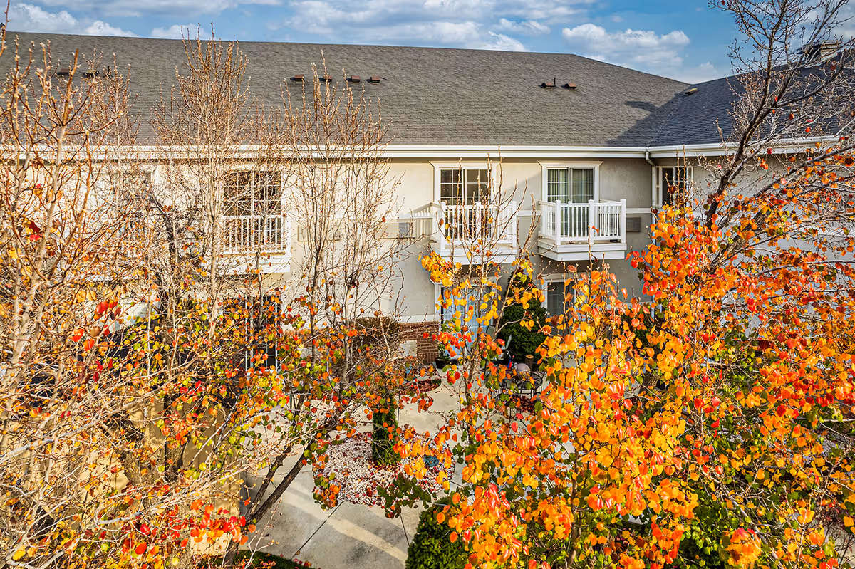 Exterior view of a two-story building with white balconies overlooking a courtyard framed by trees with bright orange and red autumn leaves.
