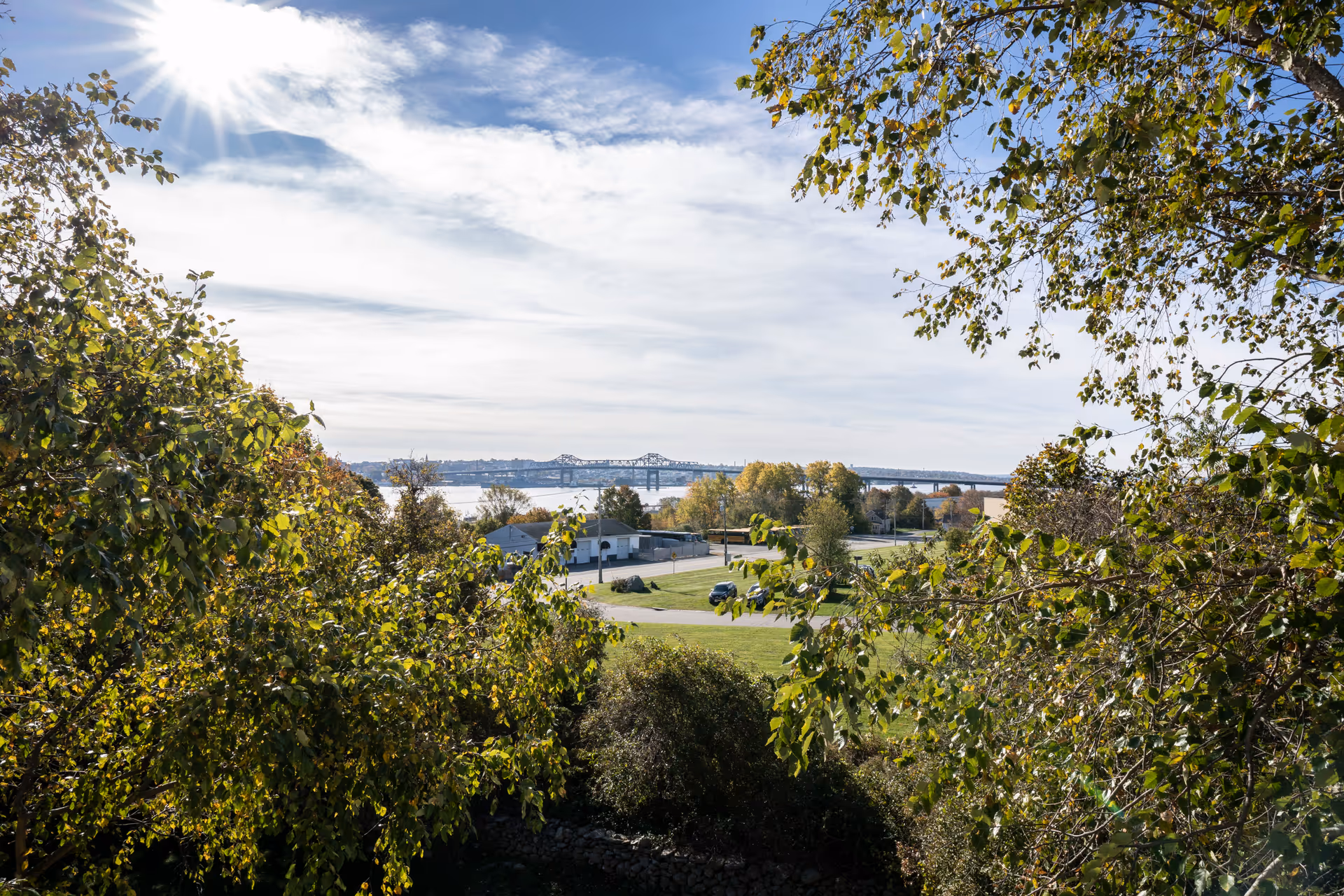 Scenic outdoor view with green trees in the foreground, a grassy area, a road, buildings, and a large bridge over a body of water under a partly cloudy sky with the sun shining brightly.