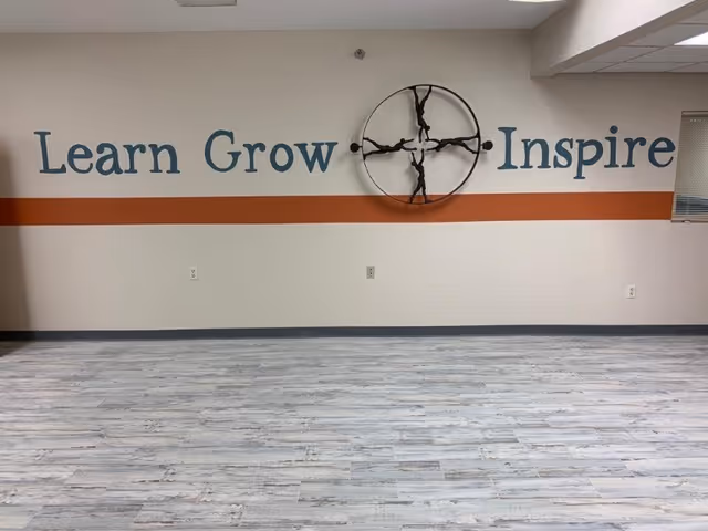 An empty room with light-colored wood flooring and a beige wall featuring a horizontal orange stripe. On the wall, the words 'Learn Grow Inspire' are painted in blue, with a decorative circular metal sculpture of four human figures in the center.