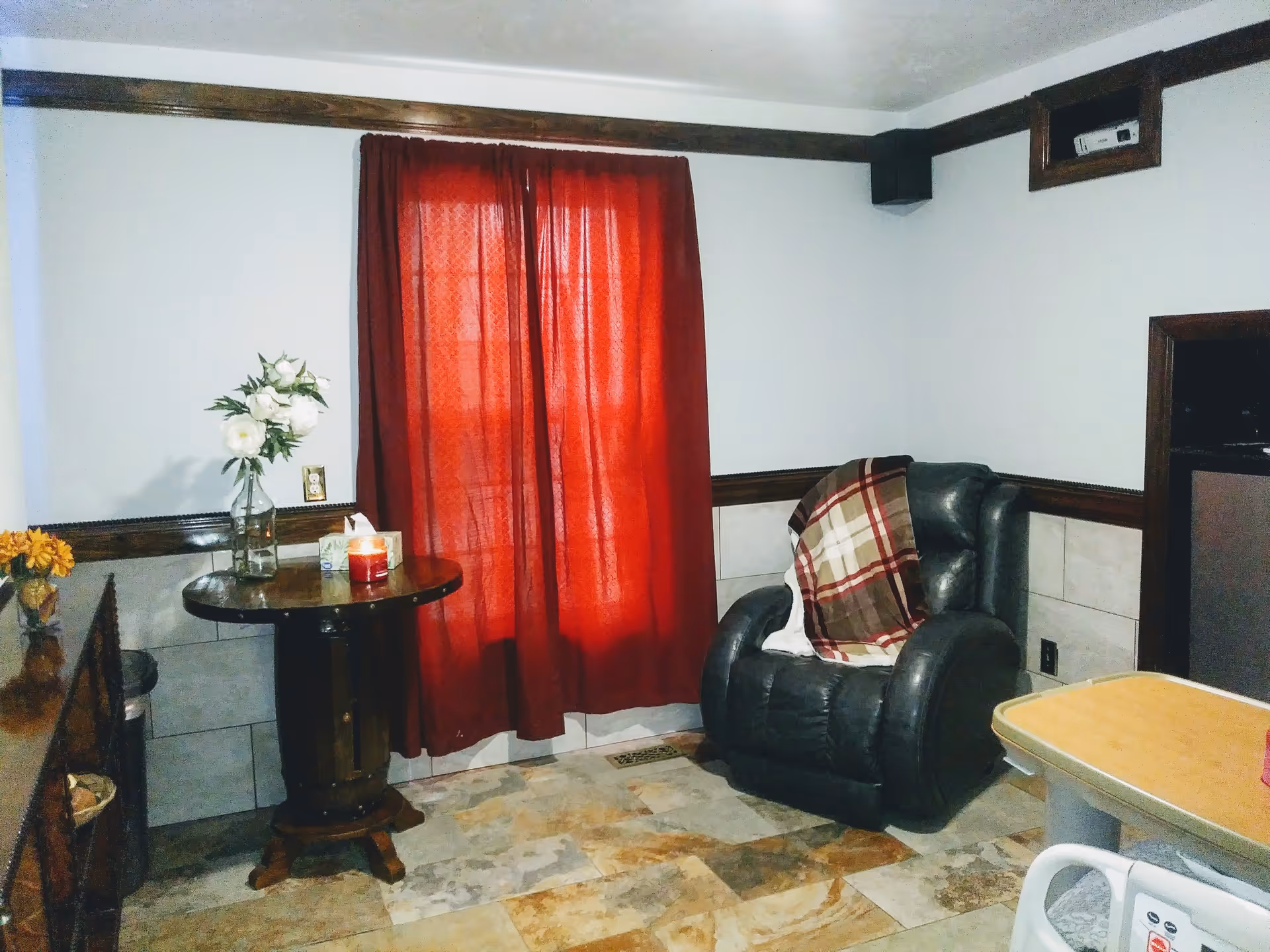 A cozy interior room with a black leather recliner chair draped with a plaid blanket, a small round wooden table holding a vase with white flowers, a candle, and a tissue box. The room has red curtains covering a window, light-colored tiled flooring, and white walls with dark wood trim.