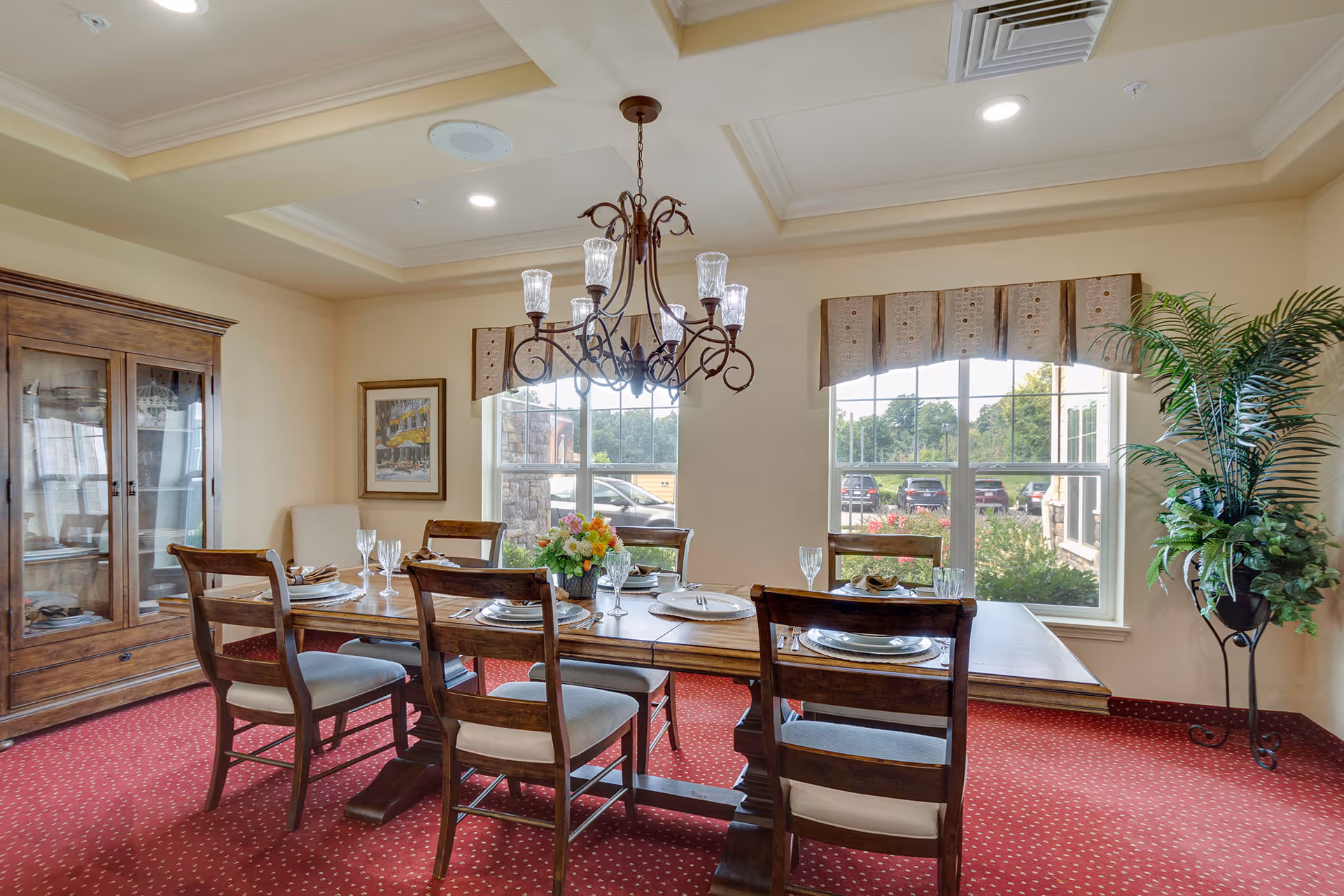 A dining room with a wooden table set for six people with plates, glasses, and napkins. The room has a red carpet with a small pattern, a wooden cabinet with glass doors on the left, two large windows with valances letting in natural light, a chandelier hanging above the table, a framed picture on the wall, and a tall potted plant on the right.
