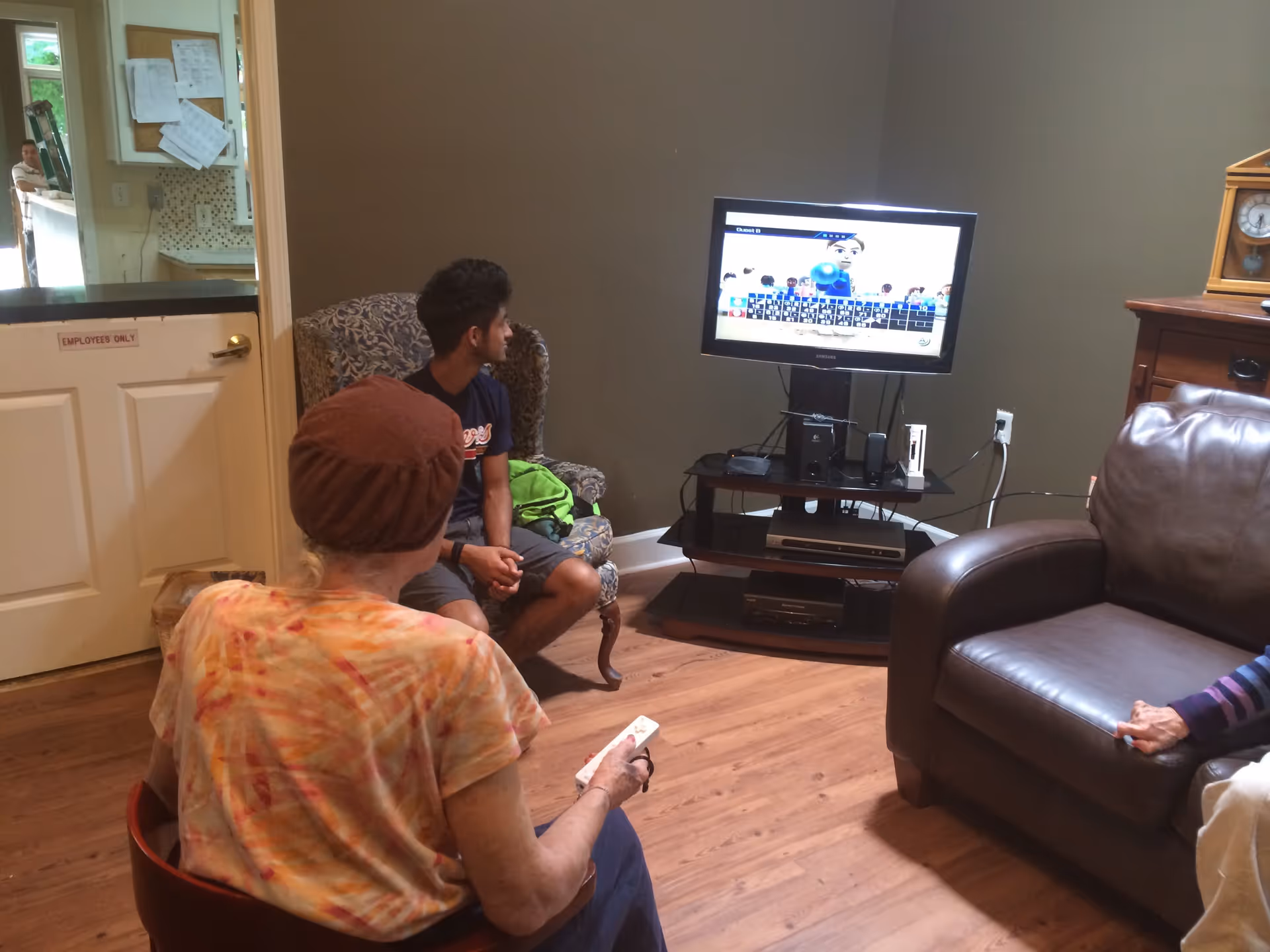 A living room with three people watching a video game on a flat-screen TV mounted on a stand. One person is sitting in a chair holding a game controller, another person is seated in an armchair, and a third person is partially visible sitting on a leather couch. The room has wooden flooring and neutral-colored walls.