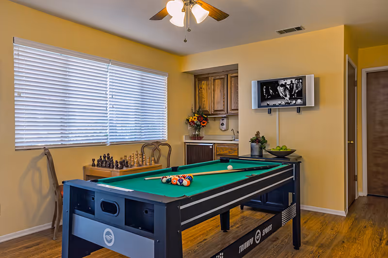 A recreational room with a pool table in the center, a chess set on a small table by the window with closed blinds, a wall-mounted TV showing a black and white image, wooden cabinets with a sink, and a ceiling fan with lights. The room has yellow walls and wooden flooring.