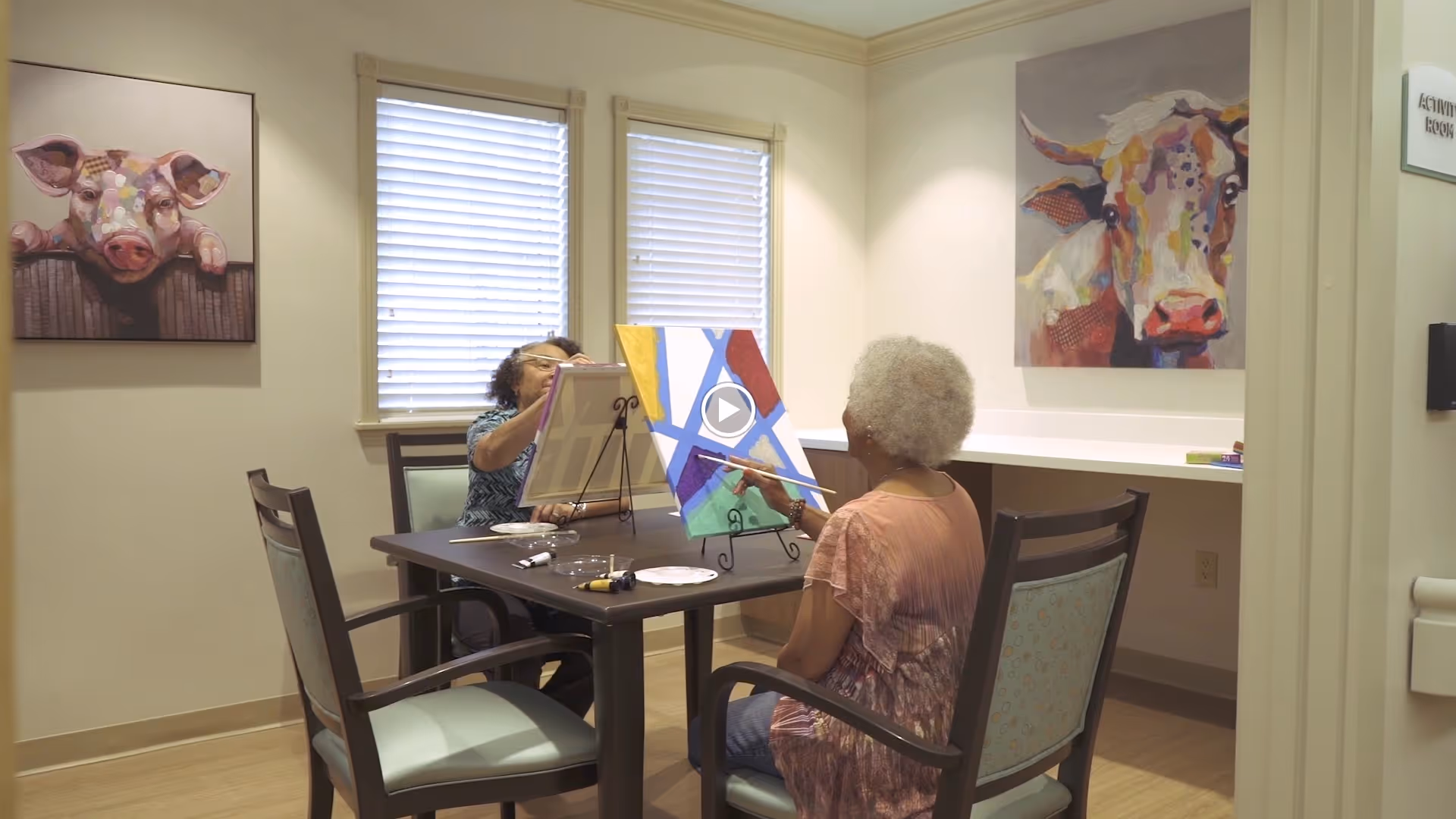 Two elderly women seated at a table in a well-lit room, engaged in painting on canvases placed on easels. The room has light-colored walls with two colorful animal paintings, one of a pig and one of a cow, hanging on the walls. There are two windows with blinds behind one woman, and the table has painting supplies on it.