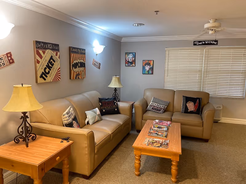 A cozy living room with two beige leather sofas, a wooden coffee table with magazines on it, and a wooden side table with a lamp. The walls are decorated with vintage-style movie posters and colorful pop art portraits. A window with closed blinds is visible in the background.