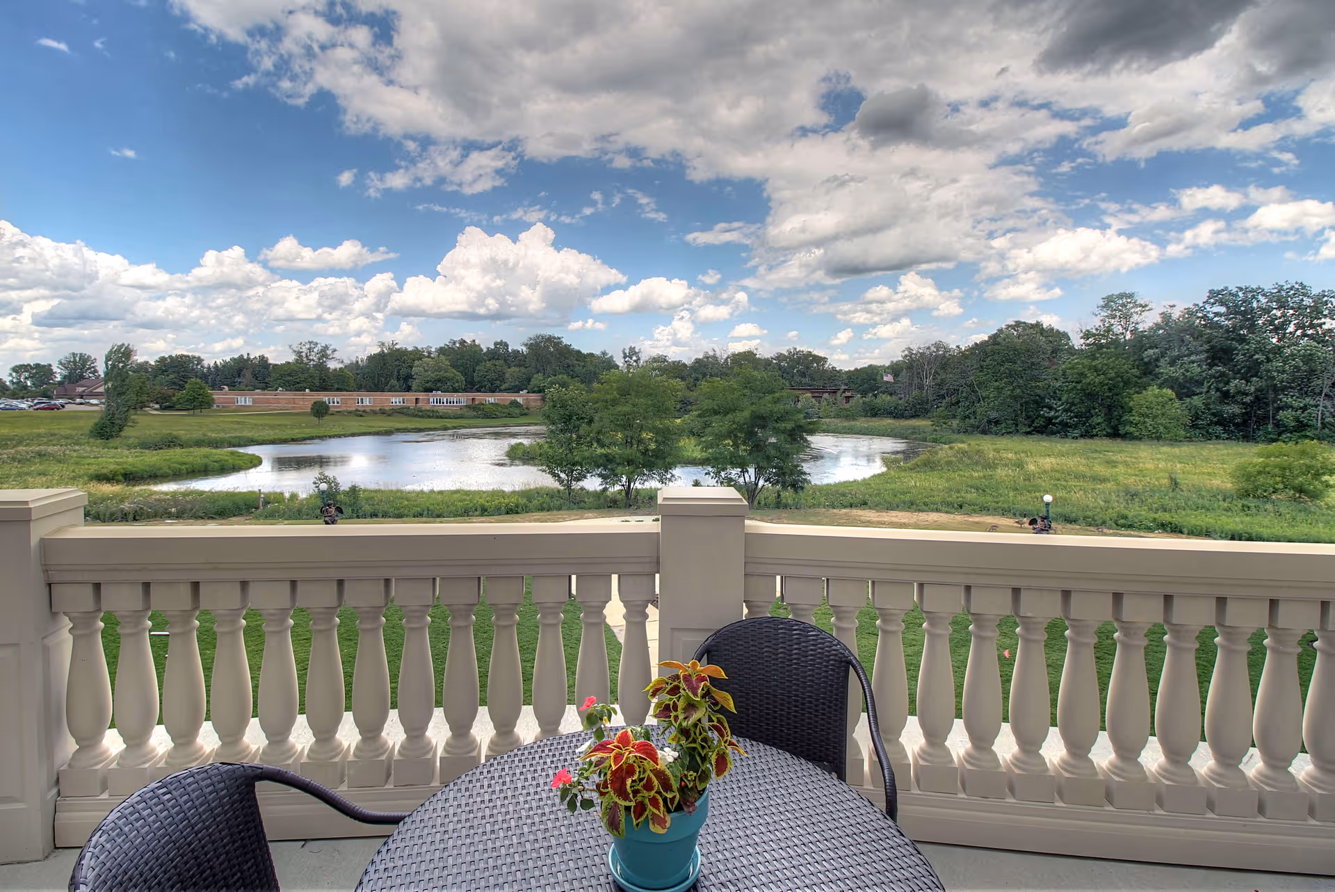 Balcony with a round table, chairs, and a potted plant overlooking a pond, grassy landscape, and cloudy sky.