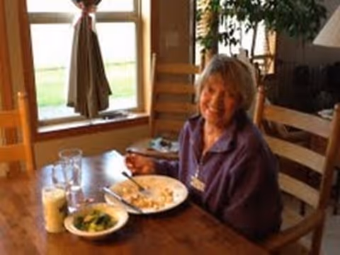 An elderly woman sitting at a wooden dining table with a plate of food, a bowl of vegetables, and a glass of milk in front of her, smiling towards the camera. The room has a window with curtains and wooden chairs around the table.