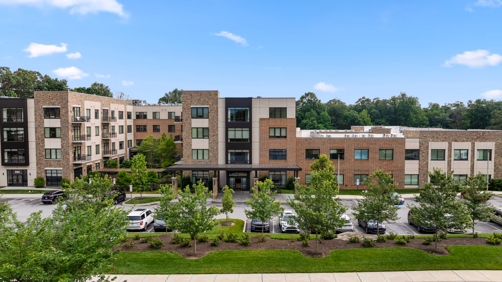 Exterior view of a modern multi-story senior living facility named Eagleview Landing, featuring a combination of brick and light-colored paneling, surrounded by trees and a parking lot with several cars parked.