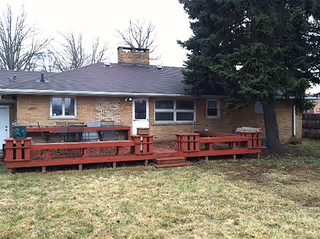 Single-story brick building with a red wooden deck and picnic-style benches facing a grassy yard and a large evergreen tree.