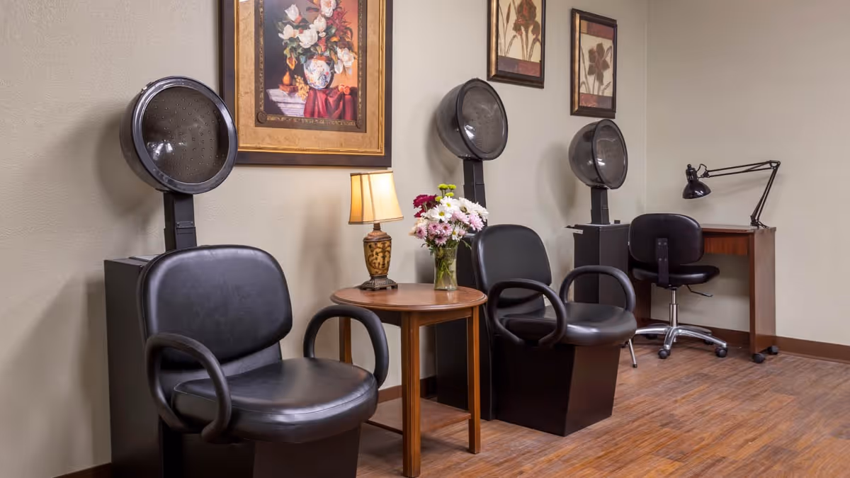 Interior view of a salon area in a senior living facility with two black salon chairs under hair dryers, a small wooden table with a lamp and a vase of flowers, and a desk with a black swivel chair and desk lamp. The walls are decorated with framed floral artwork.