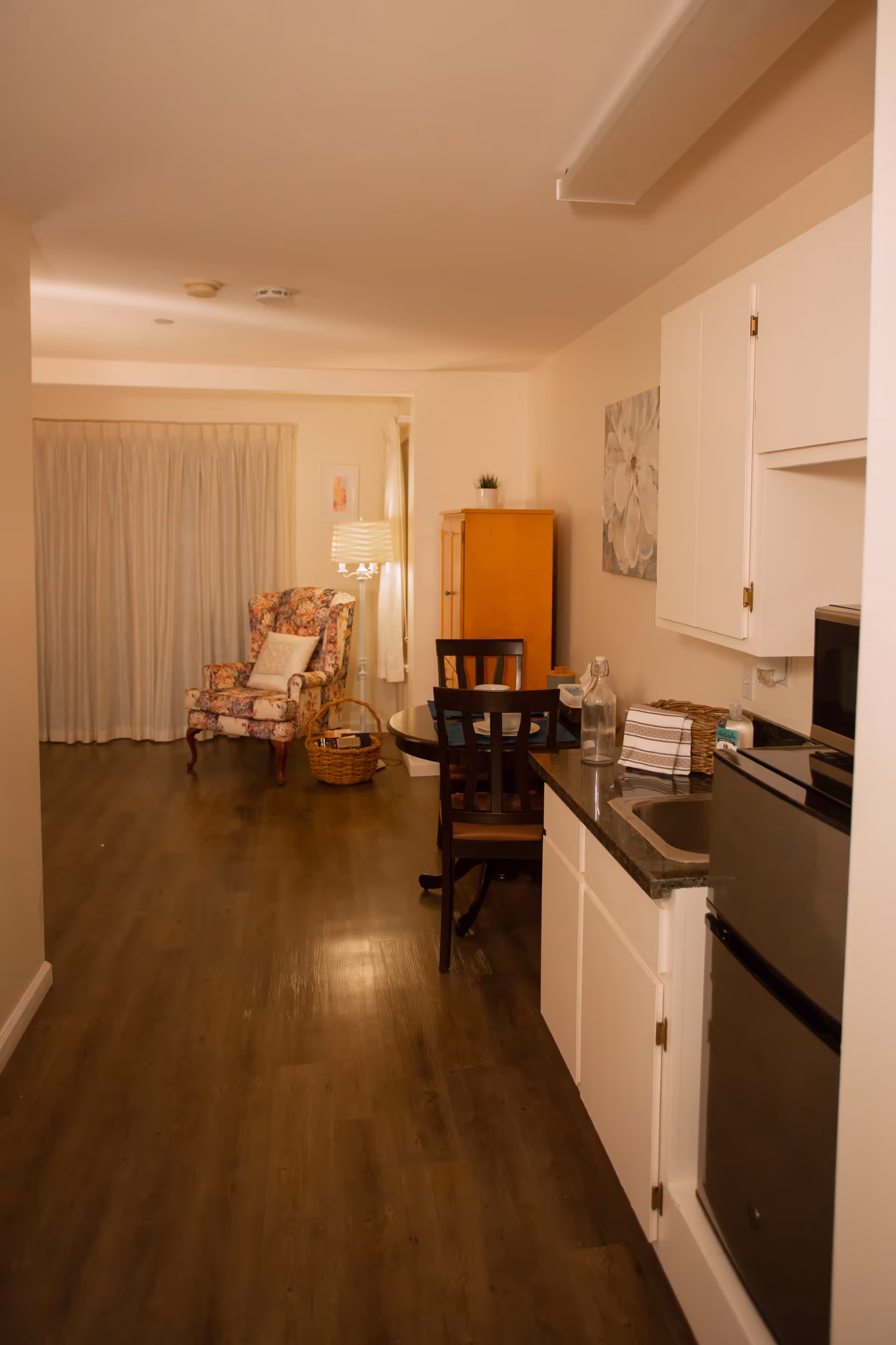 Interior view of a senior living facility room with a small kitchenette on the right, including a mini fridge, sink, and countertop. In the background, there is a round dining table with two chairs, a wooden cabinet, a floral upholstered armchair with a pillow, a basket on the floor, and a floor lamp next to a window with closed curtains.
