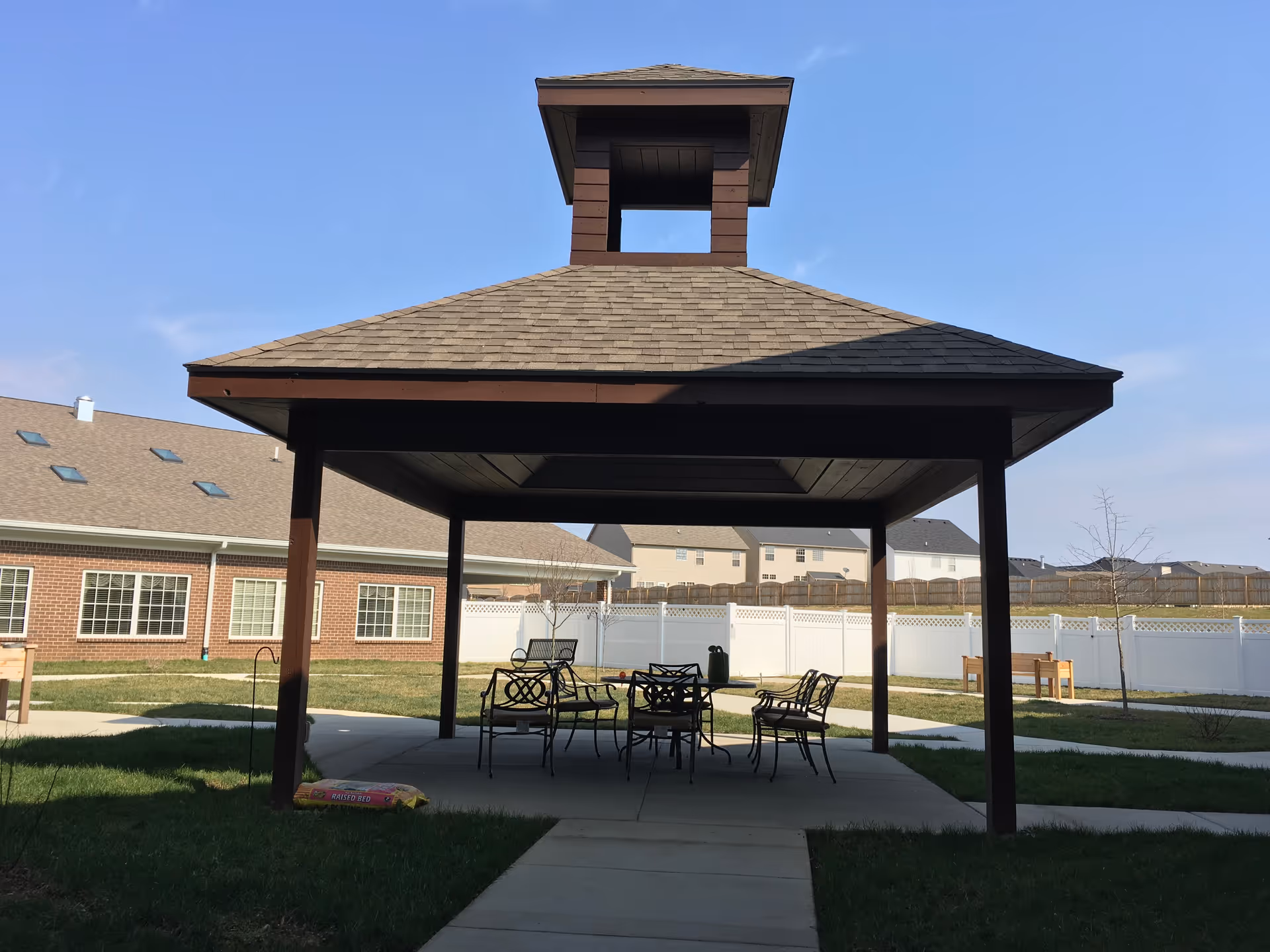 Wooden covered pavilion with a table and chairs in a fenced courtyard beside residential buildings.