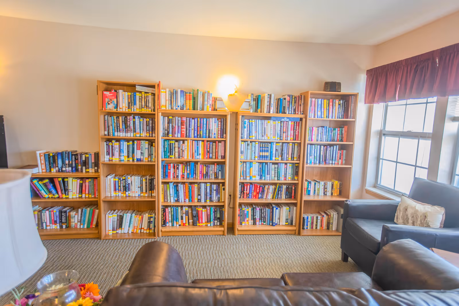 A cozy reading area in a senior living facility featuring multiple wooden bookshelves filled with books against a wall. There are two dark leather armchairs and a sofa arranged near a window with maroon curtains, creating a comfortable and inviting space.