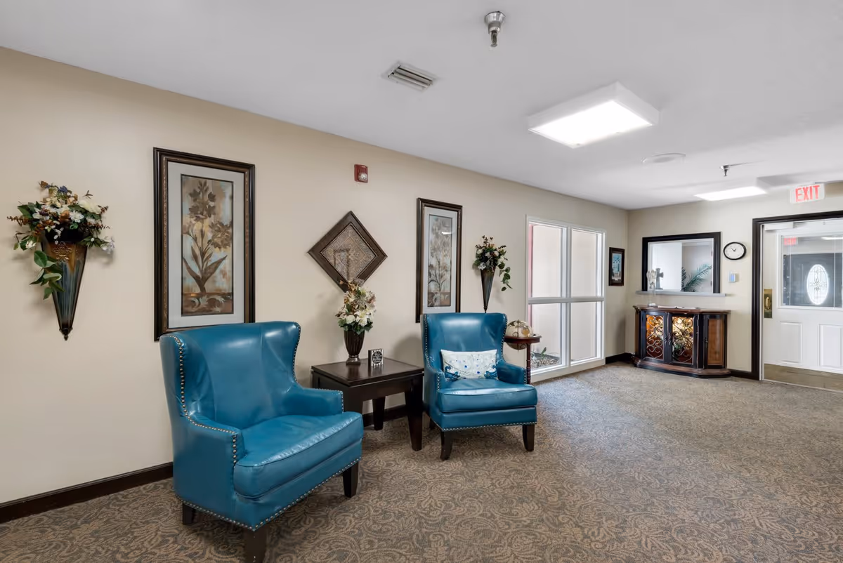 A cozy seating area in a retirement center with two blue leather armchairs, a small dark wooden table between them holding a floral arrangement, and decorative wall art and flower sconces on a beige wall. The carpet has a subtle floral pattern, and there is a window and a door with an exit sign in the background.