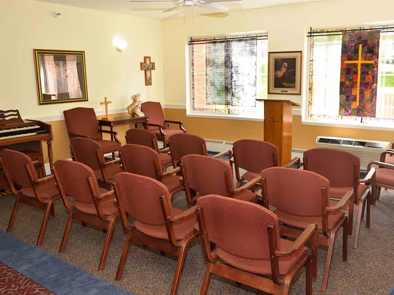 A small chapel or prayer room with rows of red cushioned wooden chairs facing a wooden podium with a cross. The room has religious decorations including a cross on the wall, a statue, a framed picture, and a stained glass window with a cross. There is also a piano on the left side of the room.