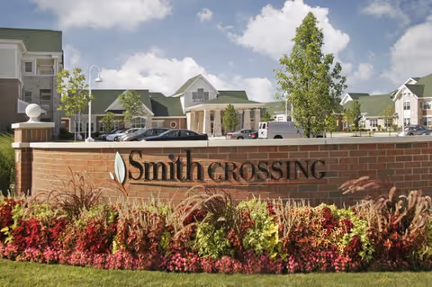 Brick entrance sign reading "Smith Crossing" with colorful landscaping and the facility buildings visible behind under a partly cloudy sky.