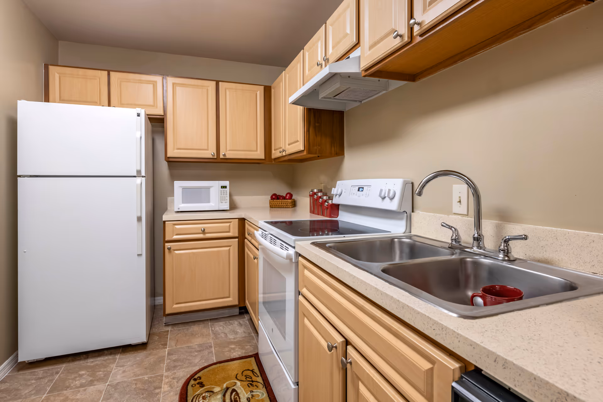 Compact kitchen with light wood cabinets, white refrigerator and stove, microwave, and a double stainless steel sink.