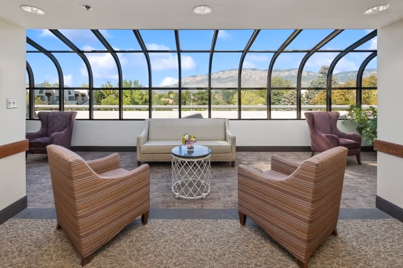 A bright indoor seating area with large curved glass windows providing a panoramic view of trees and mountains outside. The room features a beige sofa, two striped armchairs, two purple wingback chairs, and a round glass-top coffee table with a small flower arrangement.