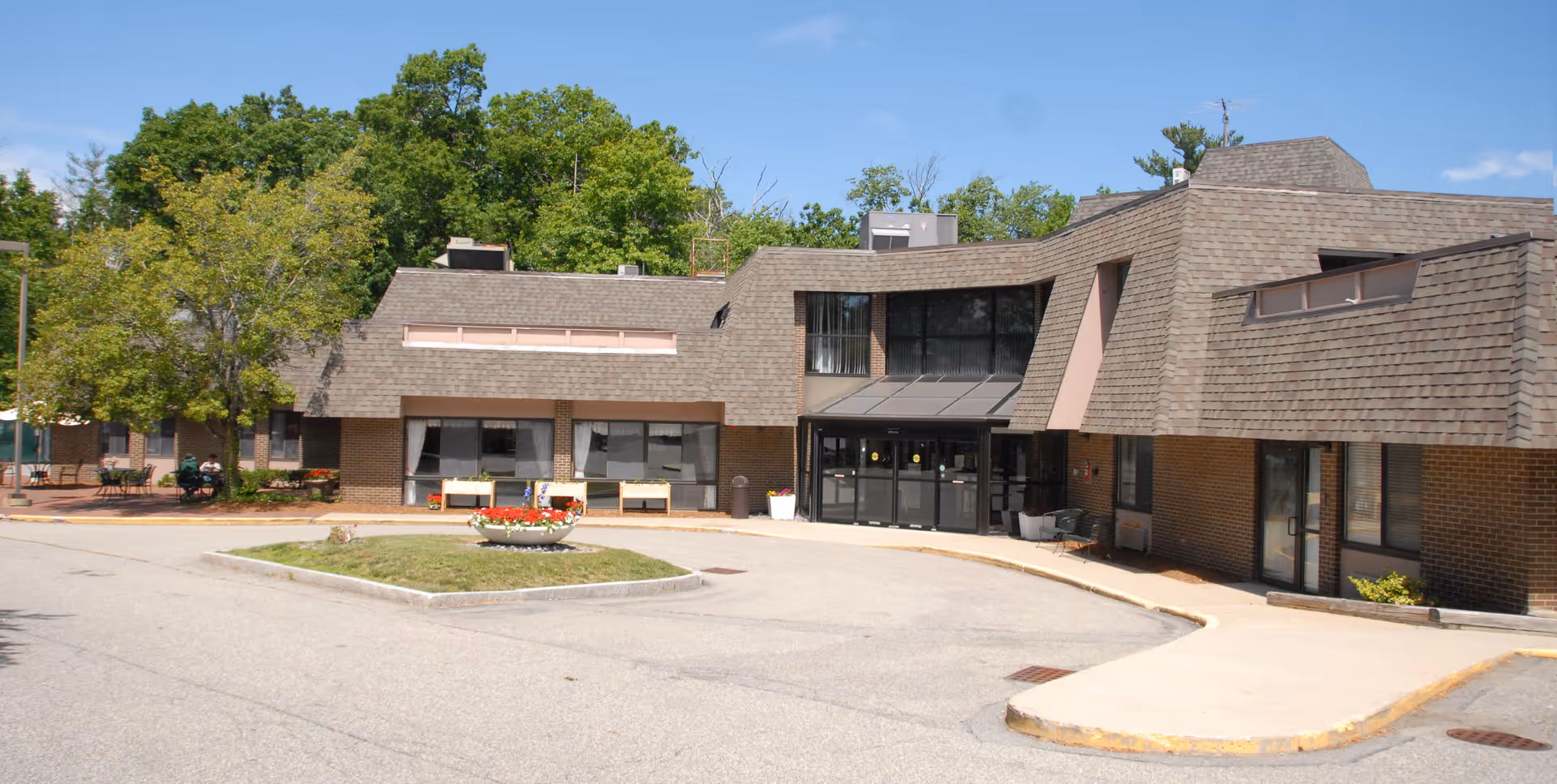 Exterior view of a senior living facility building with a brown shingled roof and brick walls. The entrance has glass doors with a small covered awning. There is a circular flower bed with red and white flowers in front of the building, surrounded by a paved driveway. Trees and greenery are visible in the background under a clear blue sky.