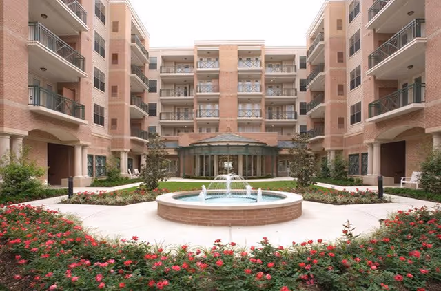 A circular fountain sits in a courtyard surrounded by red flowers and brick residential buildings.
