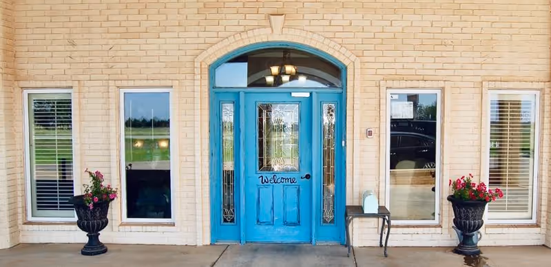 Front entrance of a building with a bright blue door that has a 'Welcome' sign on it, flanked by two tall windows on each side. There are two black planters with pink flowers on either side of the entrance, and a small bench with a white mailbox on the right side. The building exterior is made of light-colored bricks.